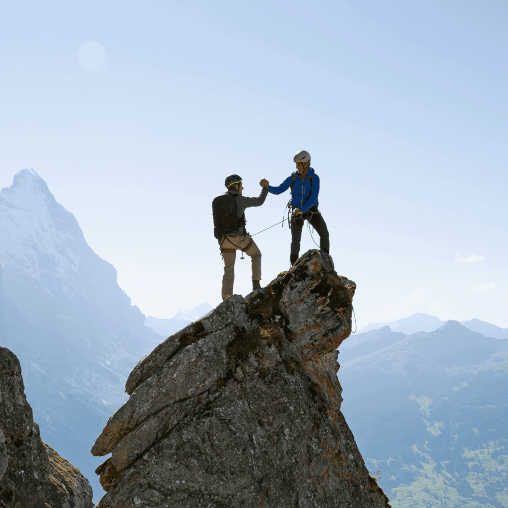 Zwei Personen stehen auf einem Berggipfel und reichen sich die Hand